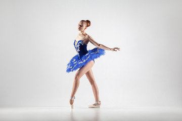 attractive ballerina with bun collected hair wearing blue dress and pointe shoes performing in white studio. © khosrork