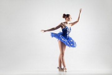 attractive ballerina with bun collected hair wearing blue dress and pointe shoes performing in white studio. © khosrork
