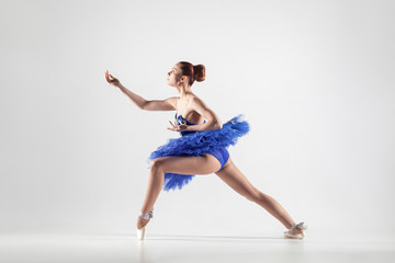 Young beautiful ballerina with bun collected hair wearing blue dress and pointe shoes dancing gracefully isolated on white background