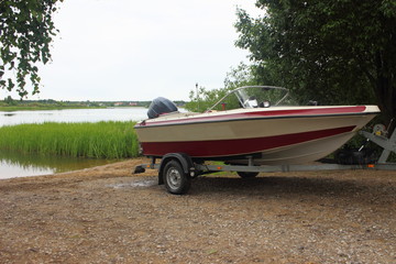 Outdoor activities - descent of a white-red motor plastic boat on a trailer with a car on the water on a sandy slipway on the background of the river and the shore with green trees