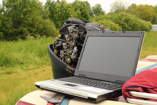 Computer diagnostics of ships - a laptop on the stern of a plastic boat on the background of a four-stroke carburetor motor, green grass and trees in summer