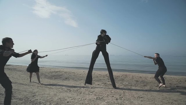 Angry mime on stilts holding the three men on leashes which are trying to topple it to the ground.