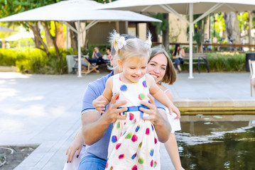 Happy laughing child girl in white dress in colored peas with her parents having fun near pond, lake in the park. Family recreation, spending time together concept. Selective focus. Copy space.