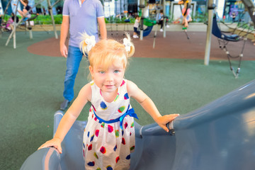 Cute little smiling blondy toddler girl in dress climbing on the childen's hill on momern summer playground with her father on the background. Selective fiocus, copy space.