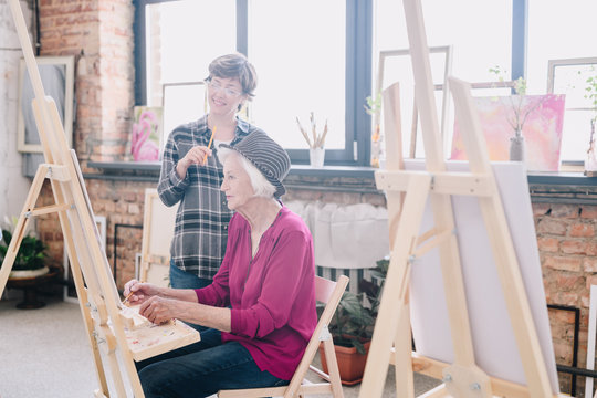 Side view portrait of elegant senior woman painting sitting at easel in art studio studying art with smiling female teacher giving comments, scene in spacious sunlit loft space.