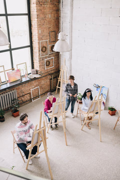 Above view portrait of adult students  painting sitting by easels in art studio with female art teacher watching them in spacious sunlit loft space, copy space