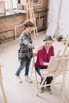 High angle portrait of elegant senior woman painting sitting at easel in art studio studying art with smiling female teacher giving comments, scene in spacious sunlit loft space.