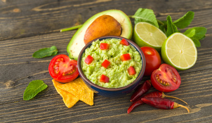 Guacamole in blue bowl on natural rustic desk with ingredient: lemon, tomatoes, peppers around.