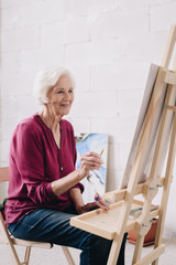 Portrait of modern senior woman painting picture sitting at easel in art studio and enjoying work in class