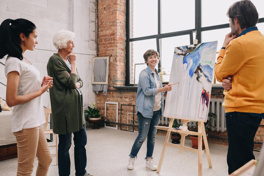 Full length portrait of female artist presenting modern watercolor painting to audience standing in art-studio or gallery
