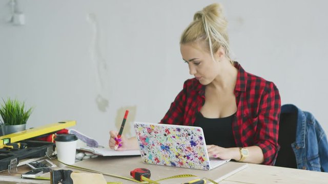 Attractive young woman in black top and checkered shirt sitting at wooden desk with tools and colorful laptop concentrating on writing in clipboard with white plastered wall on background .