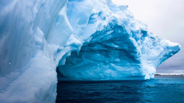 Antarctic Nature. Huge blue iceberg with natural cave inside float in open ocean. Majestic winter landscape, snow fall. Exploring, holiday, recreation, travel. Close up. 4K Slow Motion Parallax Effect