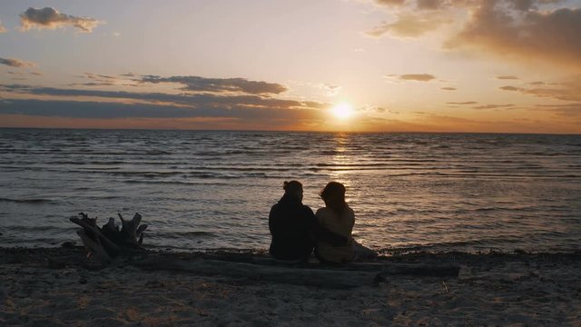A young guy and a girl are sitting on the sand by the sea, lovers are watching the waves at sunset. A loving couple is on the background of the sea. 4k