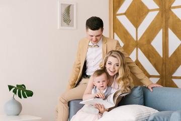 Beautiful young parents smile with their one-year-old child preview album at home in a beautiful interior in pastel colors. Family look.