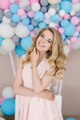 Obraz na płótnie Canvas Portrait of a beautiful young girl with curly blonde hair. Stands in a light dress against the background of white and blue balloons. Delicate hair and makeup