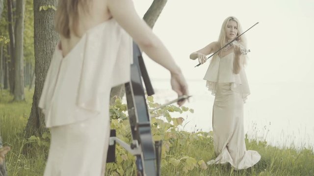 Two beautiful girls in dresses play the violin and cello standing on the edge of the sea cliff.