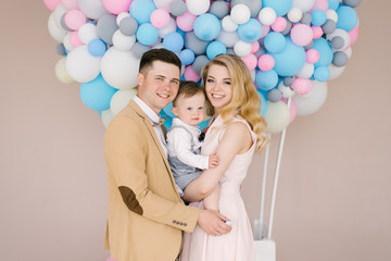 Beautiful young parents smile with their one-year-old child on pink and blue balloons background. Family look. Happy birthday party