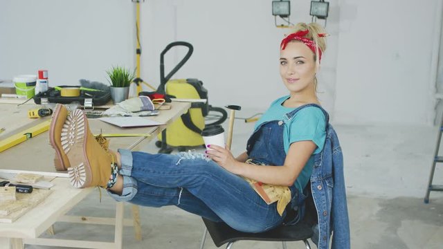 Young woman in overalls and red headband sitting comfortably on chair with legs on top of working desk full of instruments and tools holding paper cup with beverage and smiling looking at camera.