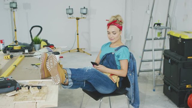Beautiful young female in jeans overalls and red headband sitting relaxed with legs on desk with tools and using mobile phone in hand