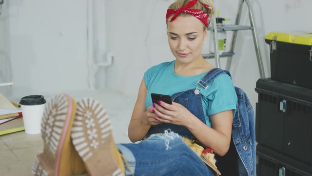 Gorgeous young female in jeans overalls concentrating on using mobile phone sitting with legs on wooden desk and tool boxes on background 