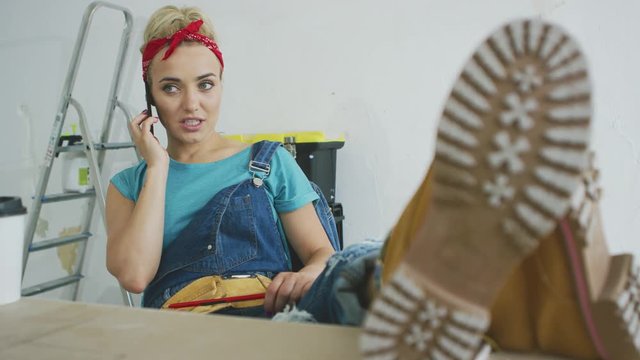 Gorgeous young blond female in jeans overalls and light work boots sitting relaxed with legs on desk talking on mobile phone and looking away over background of unpainted wall and stepladder .