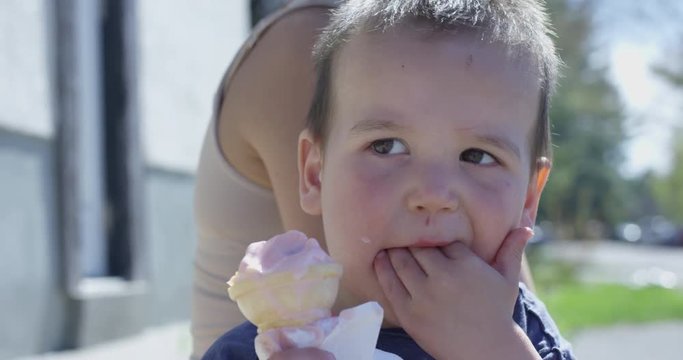Toddler boy licks fingers and puts hand in mouth while eating ice cream cone - up close