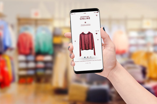 Woman hand holding smartphone and shopping online, blurred clothing store in background. Online shopping concept