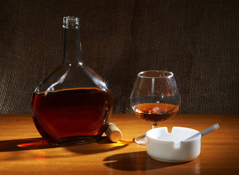 Bottle and wineglass with cognac, white ash tray and cigarette on the table with canvas background