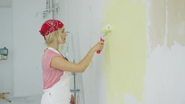 Side view of beautiful young female in white overalls and bandana using roller with pastel yellow paint on white plastered wall