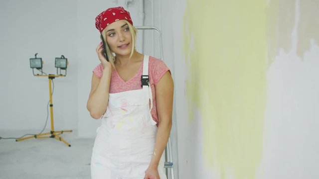 Gorgeous young female in red bandana and white overalls standing with paint roller at half-painted in pastel yellow color wall and talking on mobile phone .