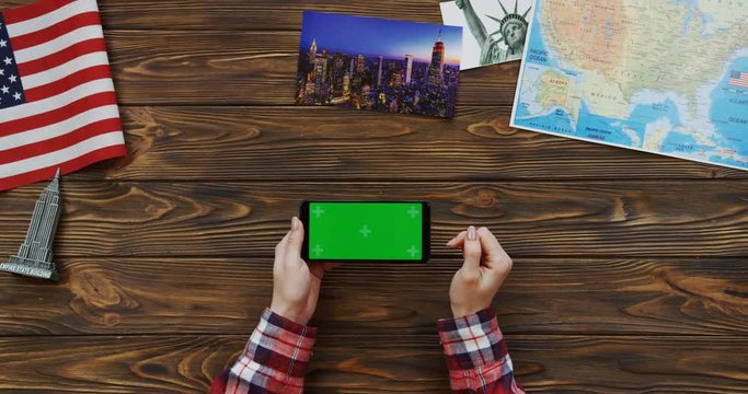 View from above on the woman's hands in plaid sleeves scrolling and taping on the horizontal black smartphone with green screen on the wooden desk with American flag, souvenirs and a map. Chroma key
