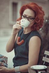 Young gorgeous redheaded woman sitting in an outdoor coffee shop, enjoying her cup of coffee, dressed in retro fashion