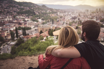Young couple in love sitting atop a hill, watching a beautiful city of Sarajevo beneath them,...