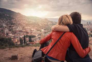 Young couple in love sitting atop a hill, watching a beautiful city of Sarajevo beneath them, enjoying a spectacular view