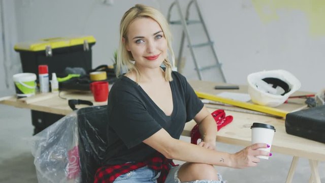 Attractive young blond female in black shirt and stylish jeans sitting comfortably with paper cup at carpenter workbench with tools and safety equipment smiling and looking at camera .