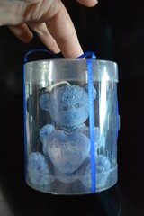 On a black glossy background is a blue candle in the form of a teddy bear in a package with a blue...