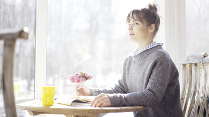 A smiling cute young brunette girl in a cafe sitting near the window dressed in a blue pullover is looking at somebody to her left in the cafe holding a pencil.