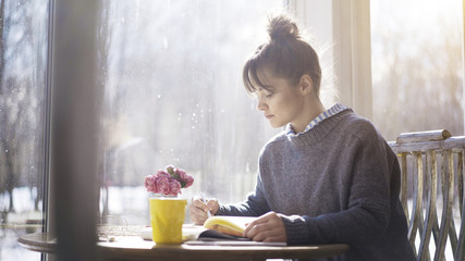 A young serious brunette girl dressed in a blue pullover is writing down notes to a notebook in the cafe sitting near the window