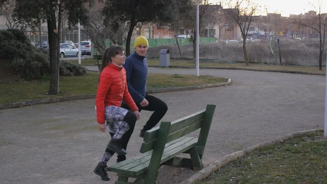 Workout with personal trainer outdoors. Young attractive woman in red bubble jacket and male fitness coach doing step-ups together onto wooden bench in park after sunset