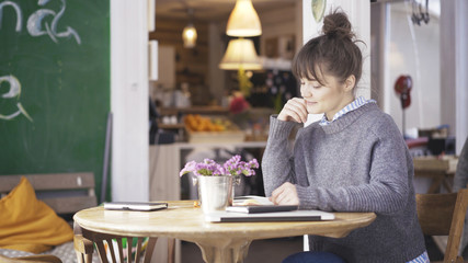 A young smiling cute brunette girl dressed in a grey pullover is reading a book in a cafe