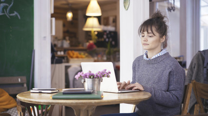Fototapeta na wymiar A young calm busy cute happy brunette girl dressed in a grey pullover is working with a laptop in a cafe