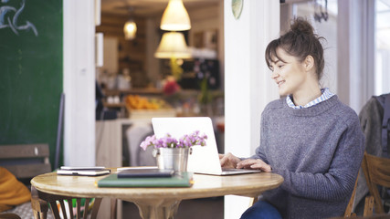 A young smiling delightful cute happy brunette girl dressed in a grey pullover is working with a laptop in a cafe