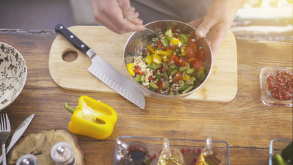 Close up of male hands mixing cucumbers with tomatoes in a metallic salad bowl with a tiny spoon