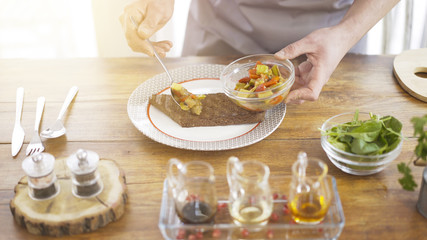A male chief is putting marinated salad on a piece of black roasted bread lying on a white plate in the kitchen