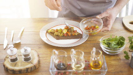 A male chief is putting marinated salad on a piece of black roasted bread lying on a white plate in the kitchen