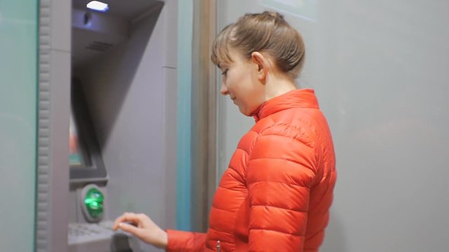 Woman using cash machine. Attractive young female in red bubble jacket inserting credit card into ATM and entering pin code