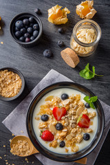 Vertical bowl of oat granola with yogurt, fresh strawberry, blueberries and nuts on black wooden table