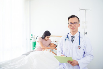 Fototapeta premium Handsome asian male doctor smiling to camera with patient and family in background