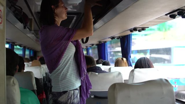 A woman walks through the cabin of the bus, removes the bag on the luggage rack and sits down