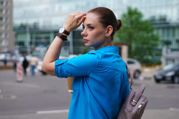 Young attractive girl with backpack holding cup of coffee. She stands on street of city. Woman adjusting her hair